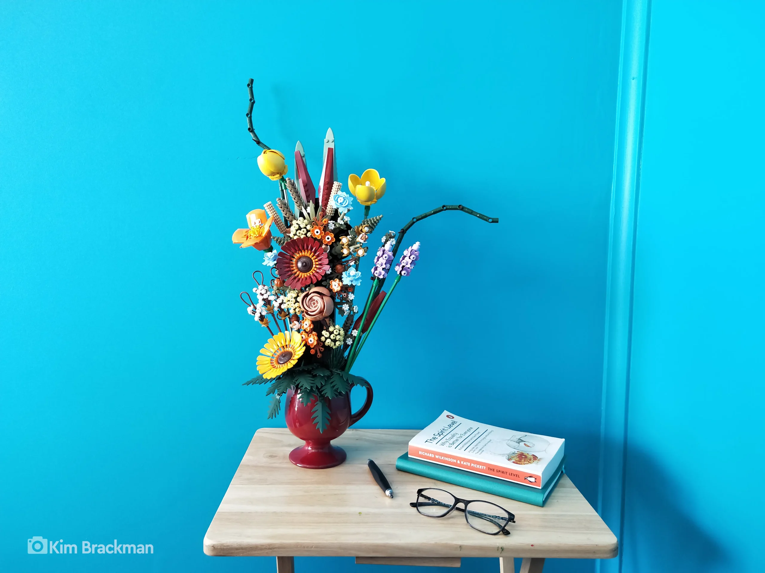 A vibrant LEGO flower arrangement in a red pitcher on a wooden table, featuring colorful flowers like sunflowers, tulips, and roses against a blue background.